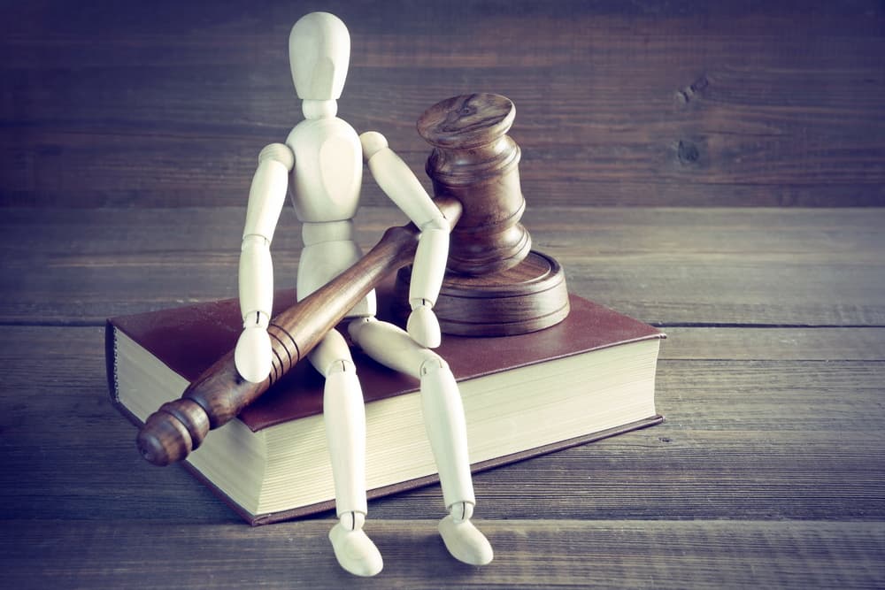 A wooden human figurine holding a judge's or auctioneer's gavel sits on a red book on a rough surface.