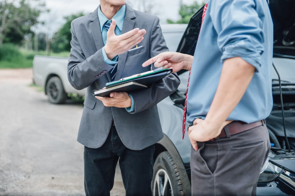 An insurance agent is processing a claim for payment from involved parties. The agent is inspecting the damaged car, and the customer is reviewing and filling out the claim report form.