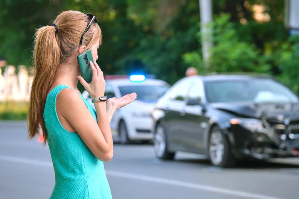 A stressed woman driver on the roadside calling emergency services on her mobile phone after a car accident. Road safety and insurance concept.