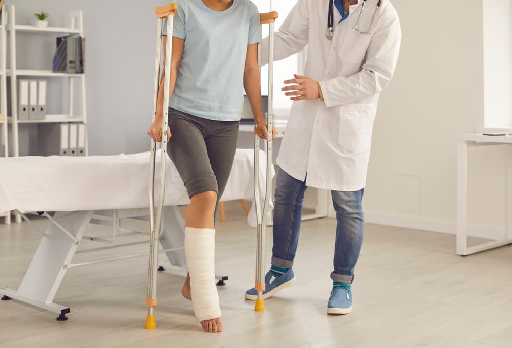 A doctor assists a patient with a broken leg. The injured young woman stands up from the examination bed at the hospital and walks using crutches.