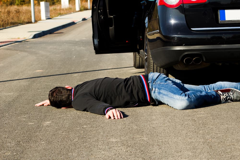 The man lies on the road behind the black car. The car collided with the pedestrian, who is now on the ground.