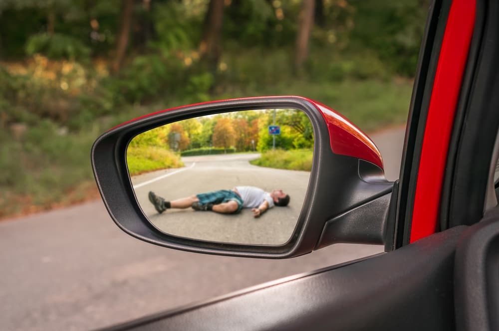 A rearview mirror reflects the scene of a car accident, showcasing a man who has been struck by a vehicle. The focus is on the mirror's perspective, creating a sense of urgency and tension.