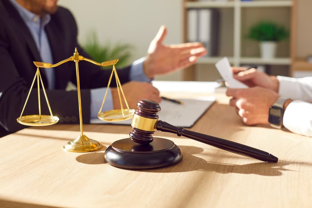 Close-up photo of a male lawyer working at his office desk, discussing a case with a male client. A judge's gavel and scales of justice are visible, symbolizing law and fairness.