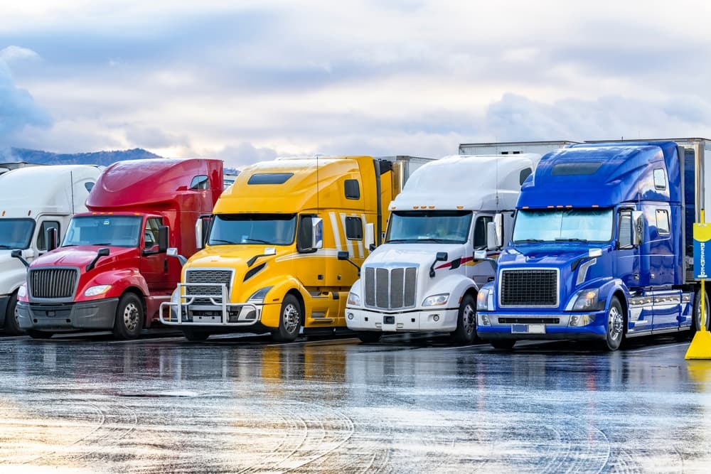 A row of industrial long-haul big rig semi trucks, each with loaded trailers, is parked at a truck stop, providing a resting area for drivers in the early morning.